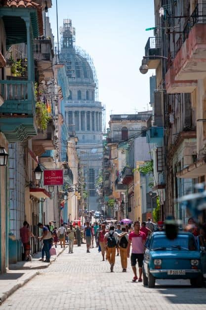 Street ambiance in Old Havana on a sunny day, with colorful buildings, old fashioned cars, cobblestoned street, and lots of people. There is a peak of the Capitolio dome in the far background. 