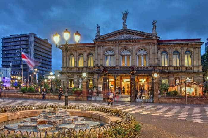 National Theater San Jose Costa Rica at night with a blue darkening sky above the impressive decorative entrance behind iron gates, the building engulfed in golden light