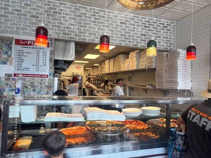 The pizza counter at Joes New York Pizza in Wynwood Miami, with lots of big pizzas displayed under warm lights, and you can see the kitchen and chefs in the background