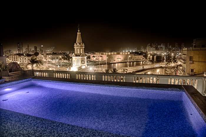 Rooftop views in Cartagena at night, with a beautiful blue pool in the foreground, and the city bathed in city lights, including the bell tower with the clock, in the background under the dark sky. 