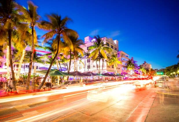 Ocean Drive in Miami Beach at night, taken with long closure time so you see the stripes of lights from cars that have passed, against the palm trees, pastel lights and restaurants in the background 