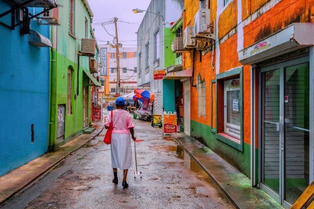 A rainy day in a narrow street in Old Havana. A woman with a stick is walking between the old houses painted in green, orange, white and blue. 