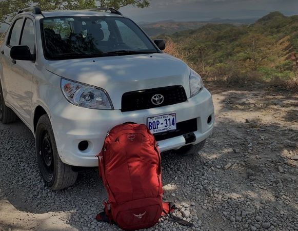 The red Osprey backpack standing beside my white rental car on a sunny day in Costa Rica. 