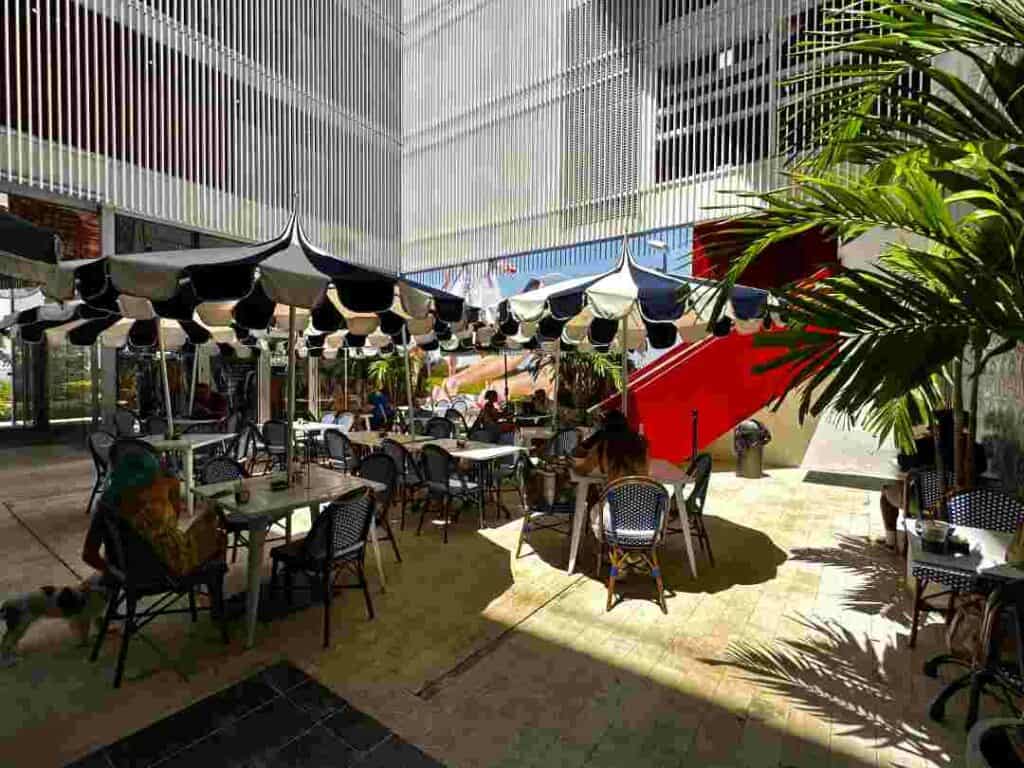 Lunch break in Brickell Miami in an open-air oasis in the middle of the forest of highrises and shopping malls. A tranquil space with green plants, seating areas and parasols, where the sun comes down between the white modern architecture surrounding the space. 
