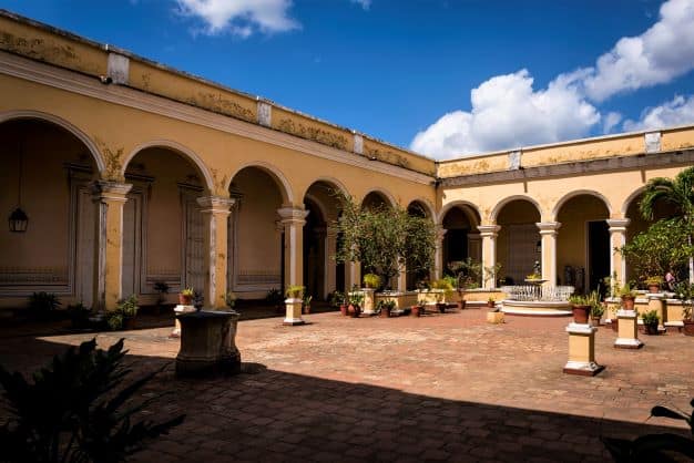 The elegant courtyard at Palace of Cantero Trinidad, a square courtyard with green bushes, yellow brick walls and shaded walkways behind elegant archways. 