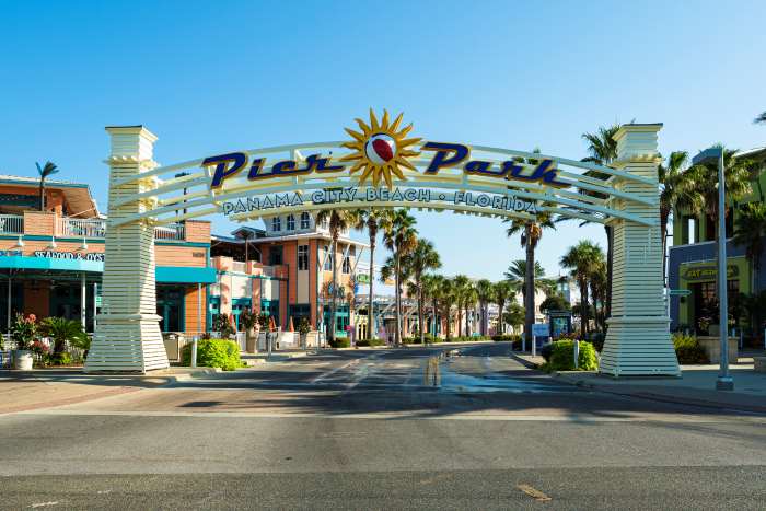 The wide white arc that signals the Panama City Beach entrance, with a colorful mall inside, and the street is lined with palm trees under the blue sky. 