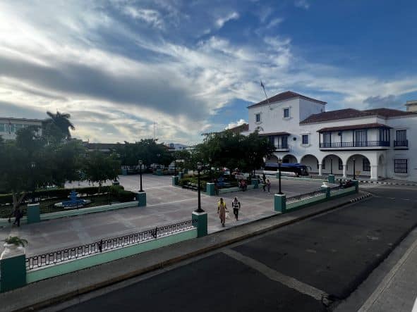 Parque Cespedes in Santiago de Cuba, a wide open square with green trees and a stone floor surrounded by white colonial houses on a partly clouded day with hazy light