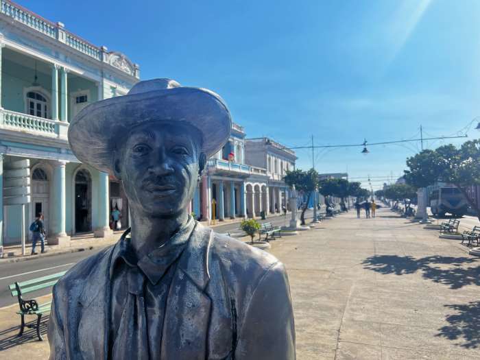 Statue on the Paseo del Prado in Cienfuegos of a full-size man with a hat, in the background the Prado disappears in the distance towards the Malecon boardwalk in the city. 