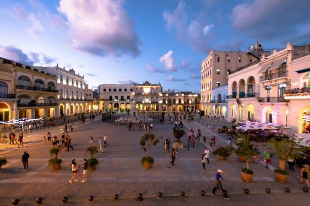 The wide Plaza Vieja in Old Havana around sunset, with lots of people enjoying the ambiance between the classic elegant buildings in a glowing light of the sunset