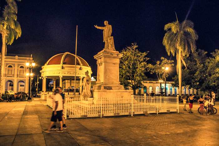 Plaza del Pueblo in Cienfuegos at night with statues, light houses, palm trees, and people walking around enjoying the evening