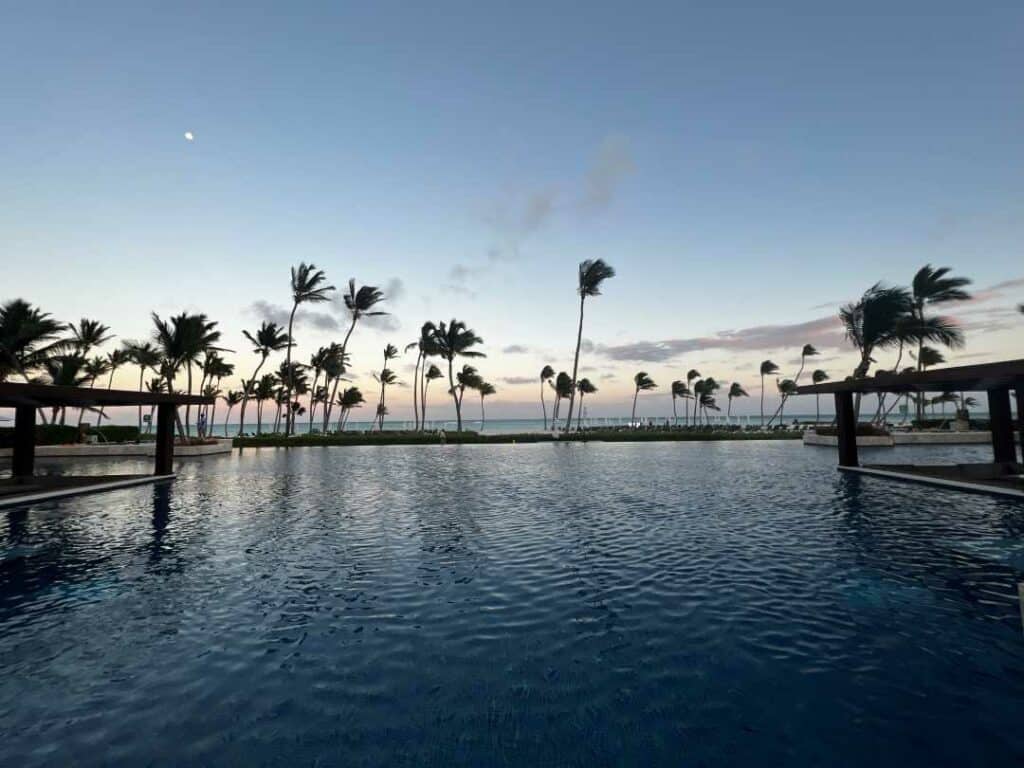 The ground level infinity pool at the Hyatt Zilara, with open views to the palm trees, beach and the ocean. On this photo it is around sunset, and the sky is pale blue with a stripe of pink near the horizon of the ocean far away. 