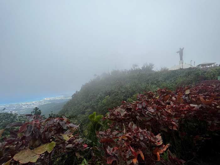 The statue of the Christ standing watch on Mount Isabel de Torres high above Puerto Plata on a day where there is fog around the mountain, but the sky is clear below us over the city and the sea. 