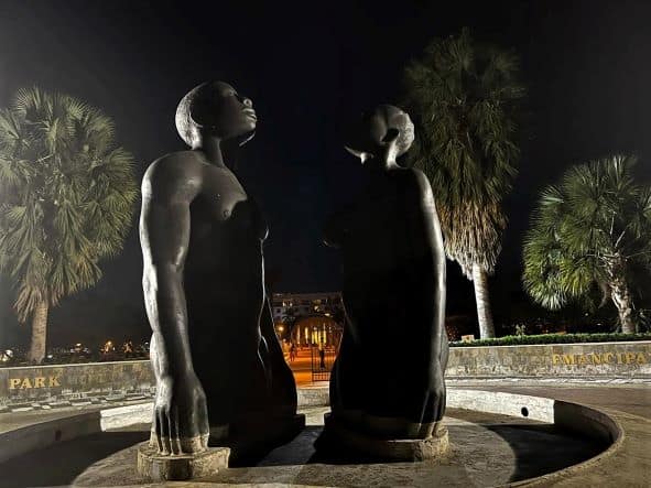 Emancipation Park in Kingston at night, a close up photo of the two naked black sculptures staring up at the sky in the park lit by strong lamps, surrounded by trees. 
