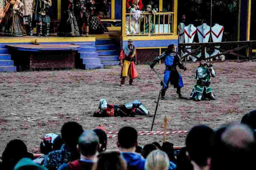 A battle ground where knights are fighting one on one in front of a stage where presumably royalty were watching from, in colorful mideaval costumes, with hundreds of spectators around 
