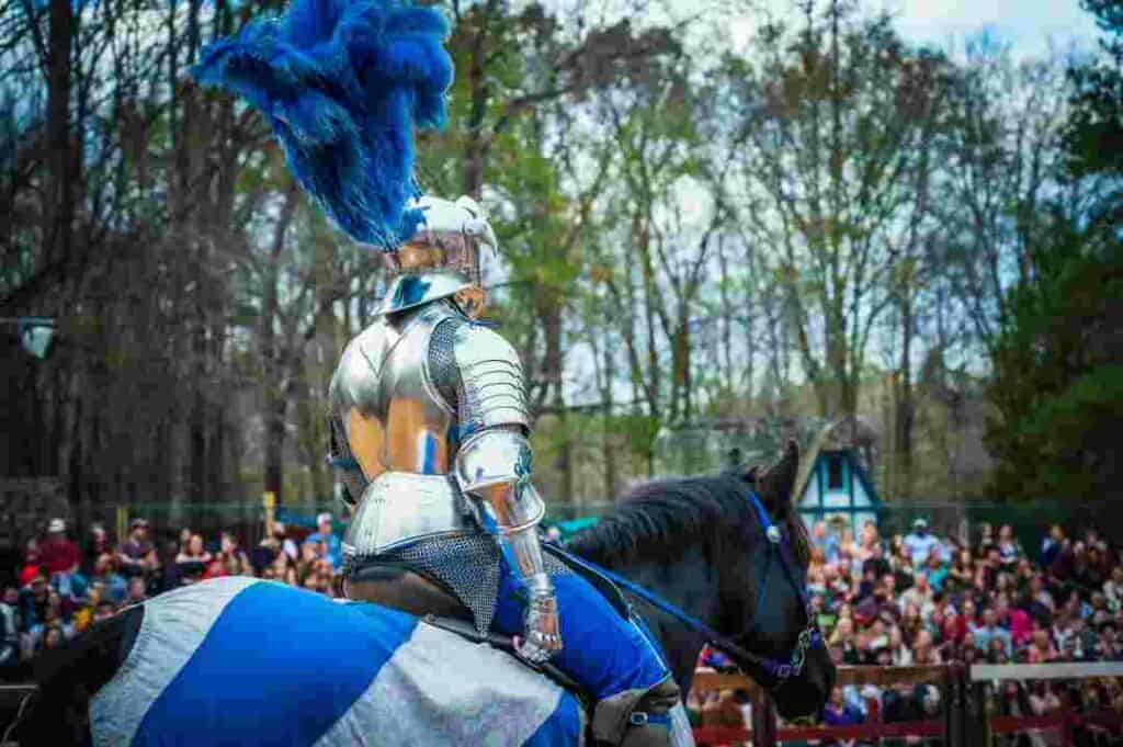 Silvery knight in his shiny armor, with bright blue feathers and accessories on horseback at a renaissance festival with hundreds of spectators