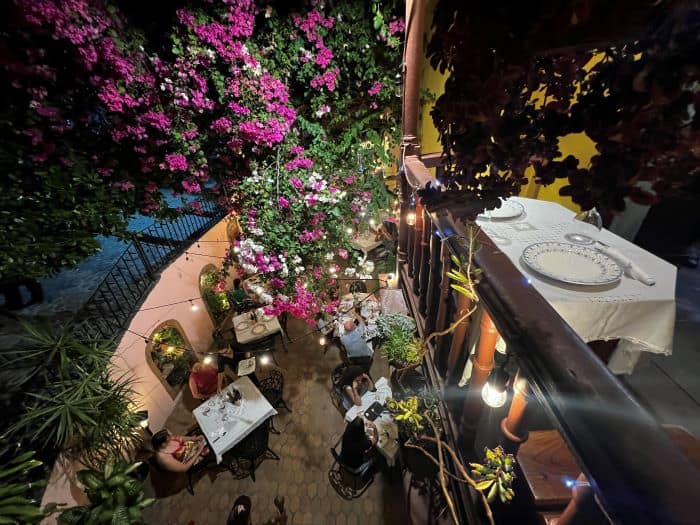 Outdoor seating at Los Conspiradores in Trinidad, a yard with charming white tables surrounded by purple and white flowers and greenery, people seated enjoying the food and ambiance at night. 