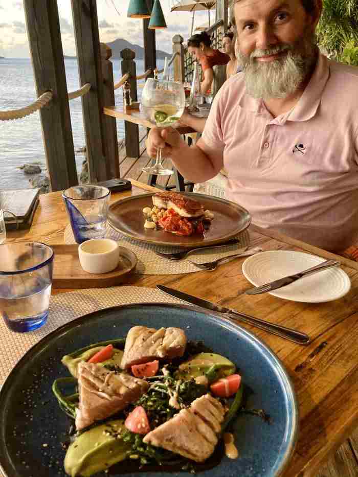 Local cuisine with a perfect ambiance, a man toasting with a glass by the outdoor table, the dinner on the plate in front of him, and a glimpse of the sea view into the horizon