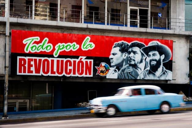 A street photo with a classic car driving past in high speed, and behind it a large red poster with pictures of heroes of the Cuban revolution, with the text "everything for the Revolution"