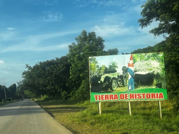 A road in rural Cuba, with a big roadside sign with a photo of the Cuban flag, a military vehicle, with the text "the land of history" in Spanish. 