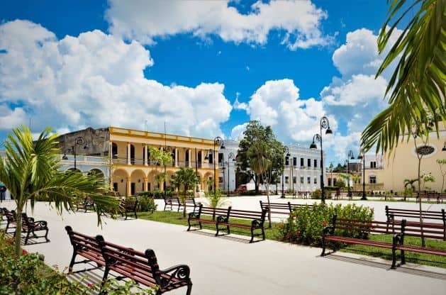 Clear bright summer day in Parque Central Santa Clara with park benches, palm trees, and classic colonial buildings in the far end. 