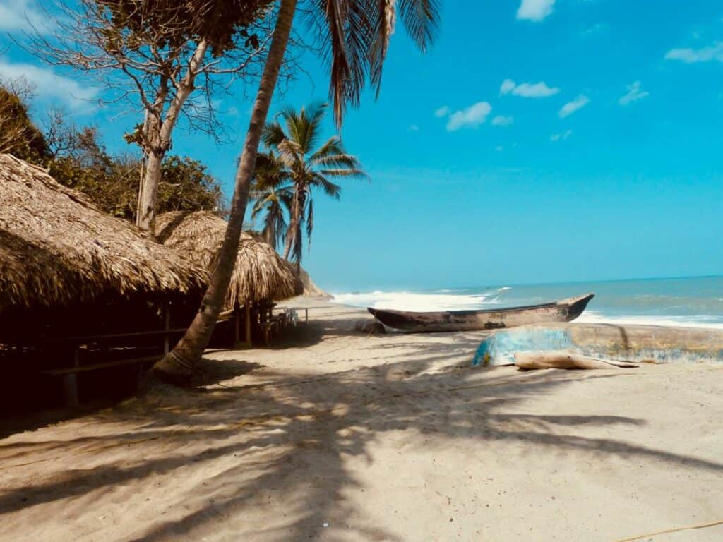 Warm sandy beaches outside Santa Marta in Colombia. Surrounded by palm trees, with a small wooden fishing boat on the shore, and traditional huts under the palm trees. 