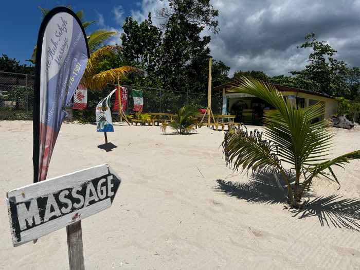 The white sands on Negril Beach in Jamaica, with a sign pointing to a beach massage parlor behind some small palmy bushes. 