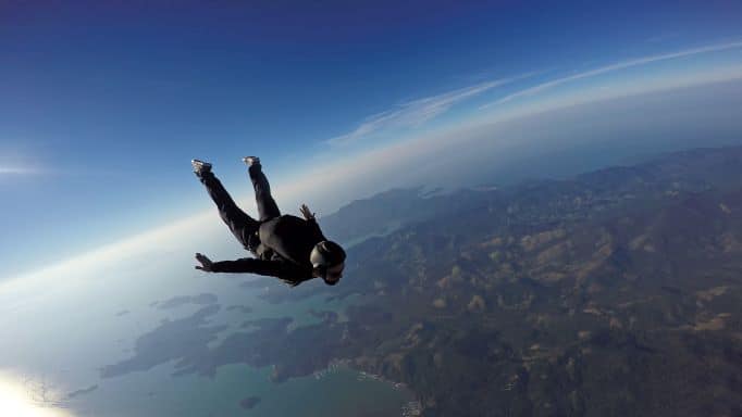 A skydiver in black flying in freefall towards the ground far below. You seem to be able to see the earth rounding far far away in the blue air in the back of the photo. 