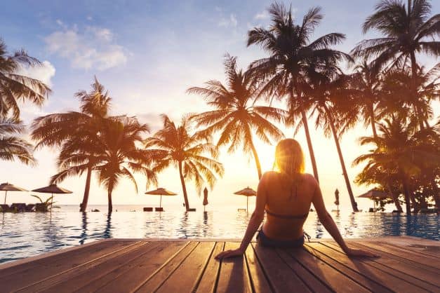 Woman sitting on a wooden jetty alone, in front of an infinity pool with palm trees and the beach on the far side, the sun is low, litght is glowing, and the sunset is not far away. 