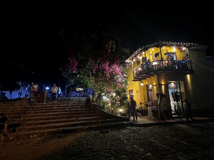 Plaza Mayor in Trinidad at night, sparingly lit from the corner restaurant in a yellow brick house with a charming balcony surrounded by bright pink flowers. 