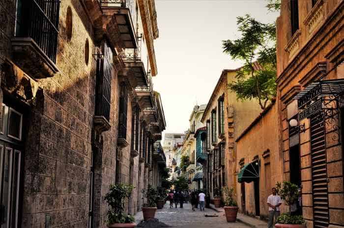 a charming street in Old Havana, with colorful stone houses lining the narrow street. The houses are painted in different warm colours or are of natural stone, and there are small balconies, intricate window decorations, and green plants in the street. 