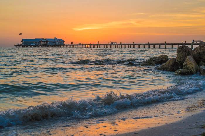 Sunrise over the Anna Maria Island pier, the waves with a bit of white foam coming onto the beach, and the pier in relieff in front of the golden sky in a distance