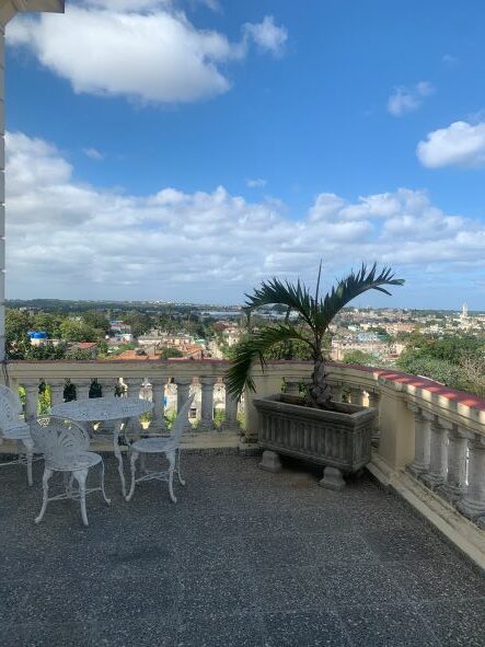 The shady terrace at Villa Teresa, with a white decorative table with chairs, green plants, and a view towards Havana. 