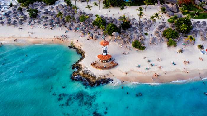 Aerial photo of the lighthouse on the sandy tip of Bayahibe Beach, with white sands meeting the light blue water, and lots of parasols tucked under the trees and bushes on shore. 