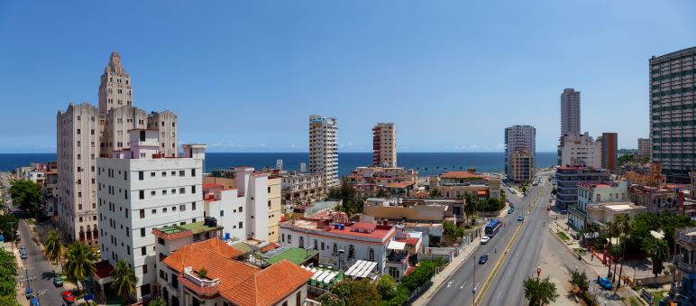 The wide streets and tall buildings in the Vedado in the day, with sun and blue skies. The sea is in hte background. 