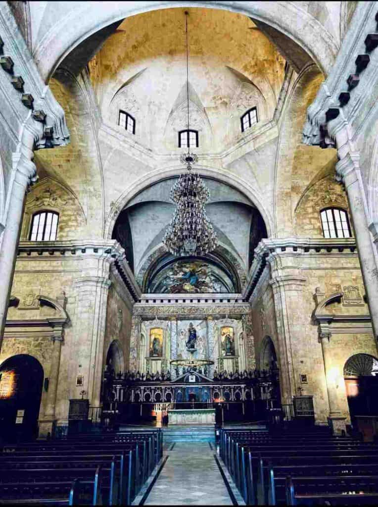 The elaborate interior of San Cristobal cathedral in Havana Cuba. From the mid section, there are dark benches on both sides, incredibly high domed ceilings, elegant structured carved out in stone, a huge chandelier, and the beautiful altar with murals and gold. 