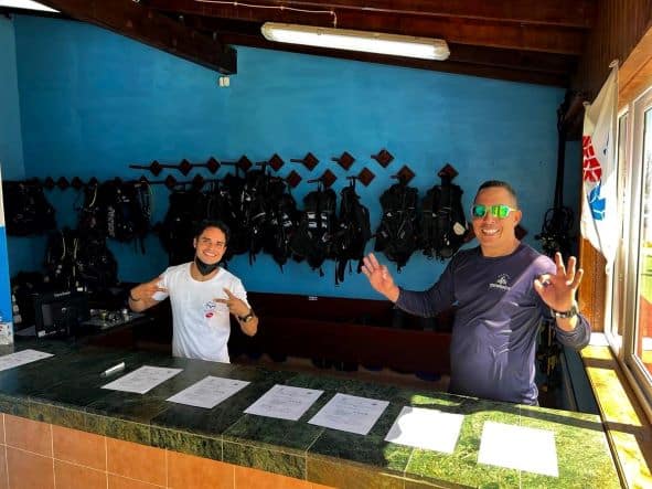 Prepping the dive gear in Marina Gaviota, from the office counter where you will get your equipment. Two happy dive guides are standing behind the counter, ready to help, and all the gear is hanging orderly in the bakcground. 