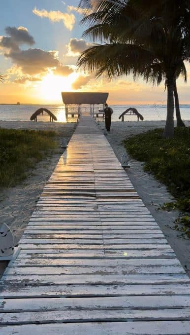 A wooden walkway leading to Sunset Beach outside the Buenavista Resort in Cayo Santa Maria. At the end of the walkway is a waiter with a glass of champagne, and behind him the stunning beach and sunset. 