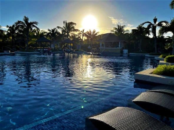 The lower pools at sunset, with elegant sun beds, lots of greenery, palm trees, and a setting sun in the distance. 