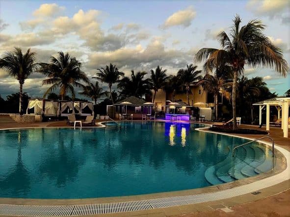 A beautiful pool in a luxury resort on Cayo Santa Maria in Cuba around sunset. Surrounded by white sun beds with pillars and curtains, palm trees, and the hotel restaurants and bar. 