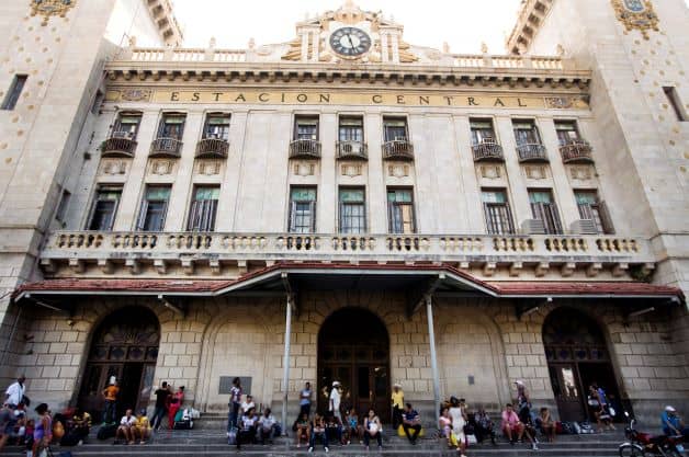 The facade of Havana train station, with lots of people sitting on the stairs outside waiting. The building is impressive, white stone with beautiful tall windows, small balconies, and elaborate details. 