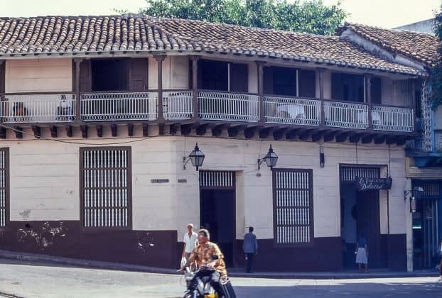 White Colonial style building in Santiago with a walkway around the second floor, brown wooden details, and a light brown stone roof. 