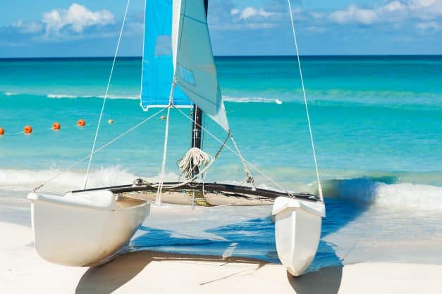 A small white catamaran with a blue sail resting in the surf on the beach in Varadero on white sands with crystal clear water coming in. 