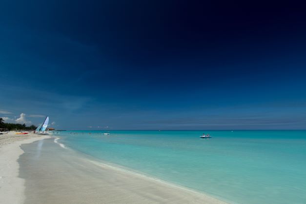 The beautiful white sandy Varadero Beach with a windsurf board in the distance along the beach. The water is light greenish and crystal clear against the deeper blue sky on a sunny summer day. 