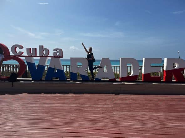 Posing in front of the large red, white and blue sign spelled Varadero, by one of the entrances to Varadero beach. The ground in the forefront is red, and behind me, there is blue sea and sky. 