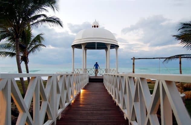 A beautiful wooden walkway to the beach in Varadero, with white railings, and you can see the greenish crystal clear water outside, and the light blue early evening sky. 