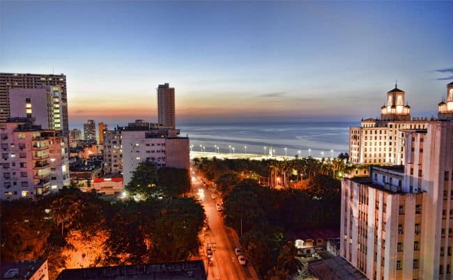 The Vedado, the modern district of Havana at night seen from above towards the sea. Streets and buildings are lit, and the sky is pale pink in the distance from the sun that has just set. 