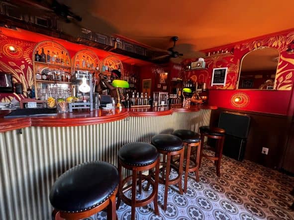 The bar at the Viajero Miami in South Beach, with brown stools with black seats next to the elegant art deco style par in reddish wood, the walls decorated with red patterned wall paler