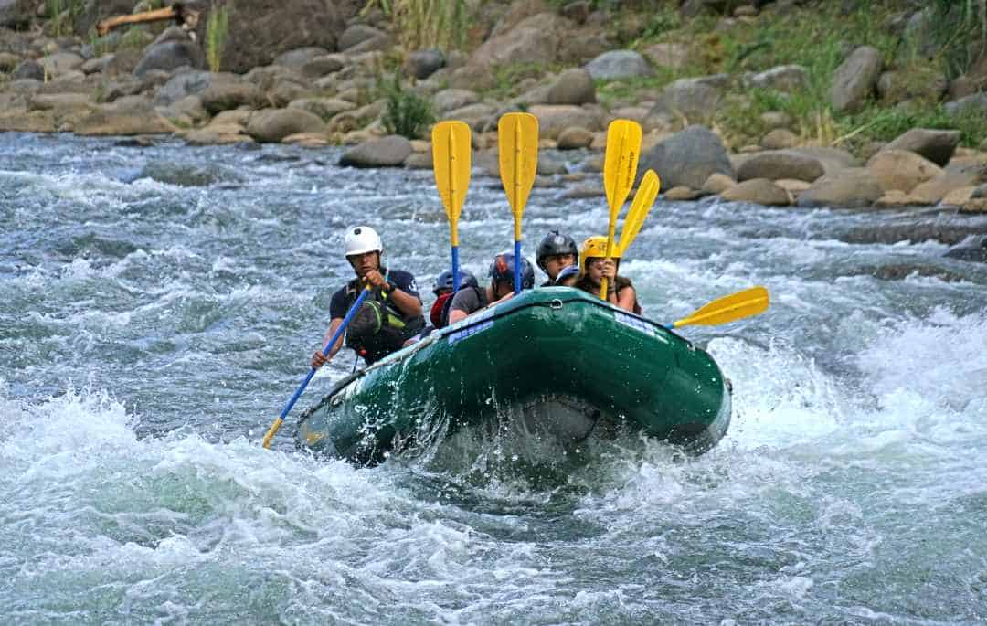 Go whitewater rafting in Costa Rica, down the wild river, like these people in a green boat, with yellow paddles, wearing helmets while racing down a fierce river