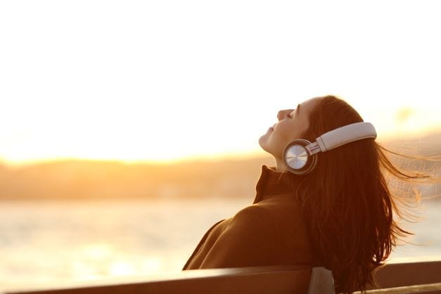 Wherever you are: relax, and breathe. A woman sitting on a bench with eyes closed, smiling, with headphones on. In the unclear distance behind her is the golden sunset. 