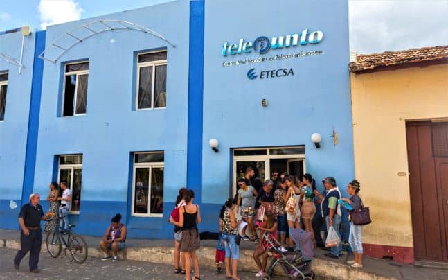 A light blue building that is home to the Etecsa telephone store in Cuba, with a line of people waiting to get in. 